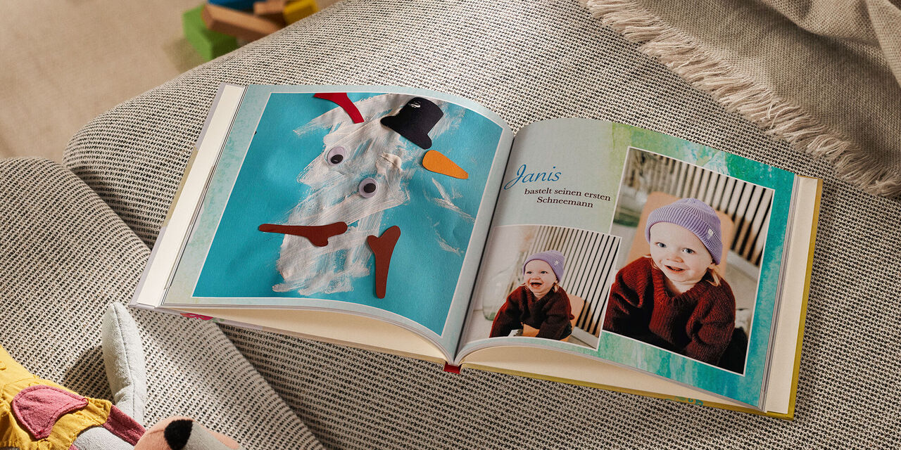 Auf einem Sofa liegt ein geöffnetes CEWE FOTOBUCH neben einem Kuscheltier und einer Decke. Die linke Seite zeigt das Foto einer Kinderbastelarbeit, die einen Schneemann darstellt. Auf der rechten Seite sind zwei Fotos von einem lachenden Kind zu sehen. Die Überschrift lautet «Janis bastelt seinen ersten Schneemann». Der Hintergrund der Doppelseite ist mit blau-grünen Aquarellfarben ausgefüllt.